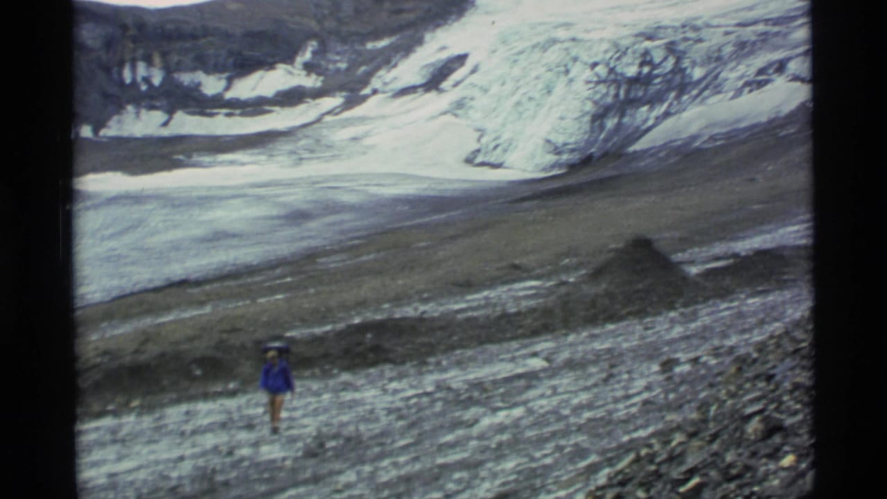Distant hiker in a blue jacket crossing rocky ground below a glacier.