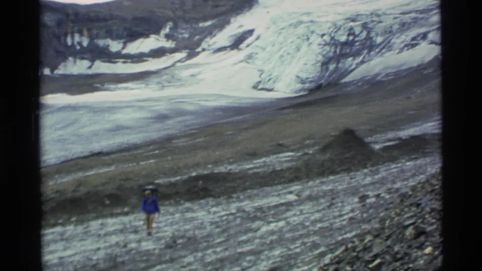 1977 Alaska 8mm frame showing a distant blue-jacketed hiker on rocky moraine beneath glacier ice and mountain slopes.