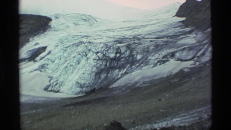1977 Alaska 8mm frame emphasizing glacier ice, dark rock bands, snow, and a barren moraine landscape.