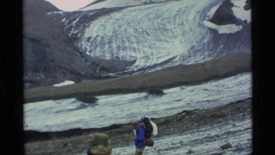 1977 Alaska 8mm frame showing a backpacked hiker in blue near snow-covered ground below a glacier and mountain slope.