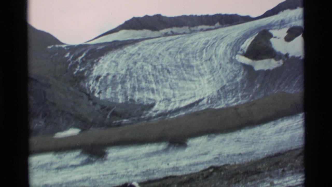 1977 Alaska 8mm frame showing a broad glacier surface, dark ridgelines, snow patches, and a band of water or moraine across the ice.