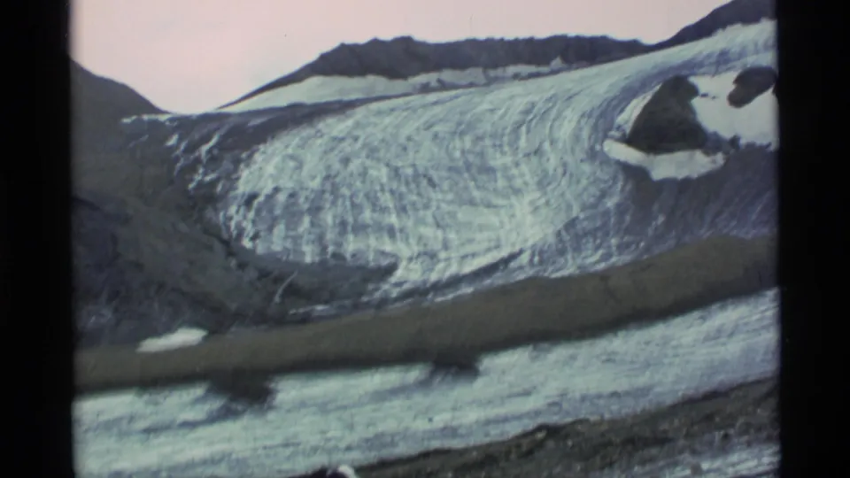1977 Alaska 8mm frame showing a broad glacier surface, dark ridgelines, snow patches, and a band of water or moraine across the ice.