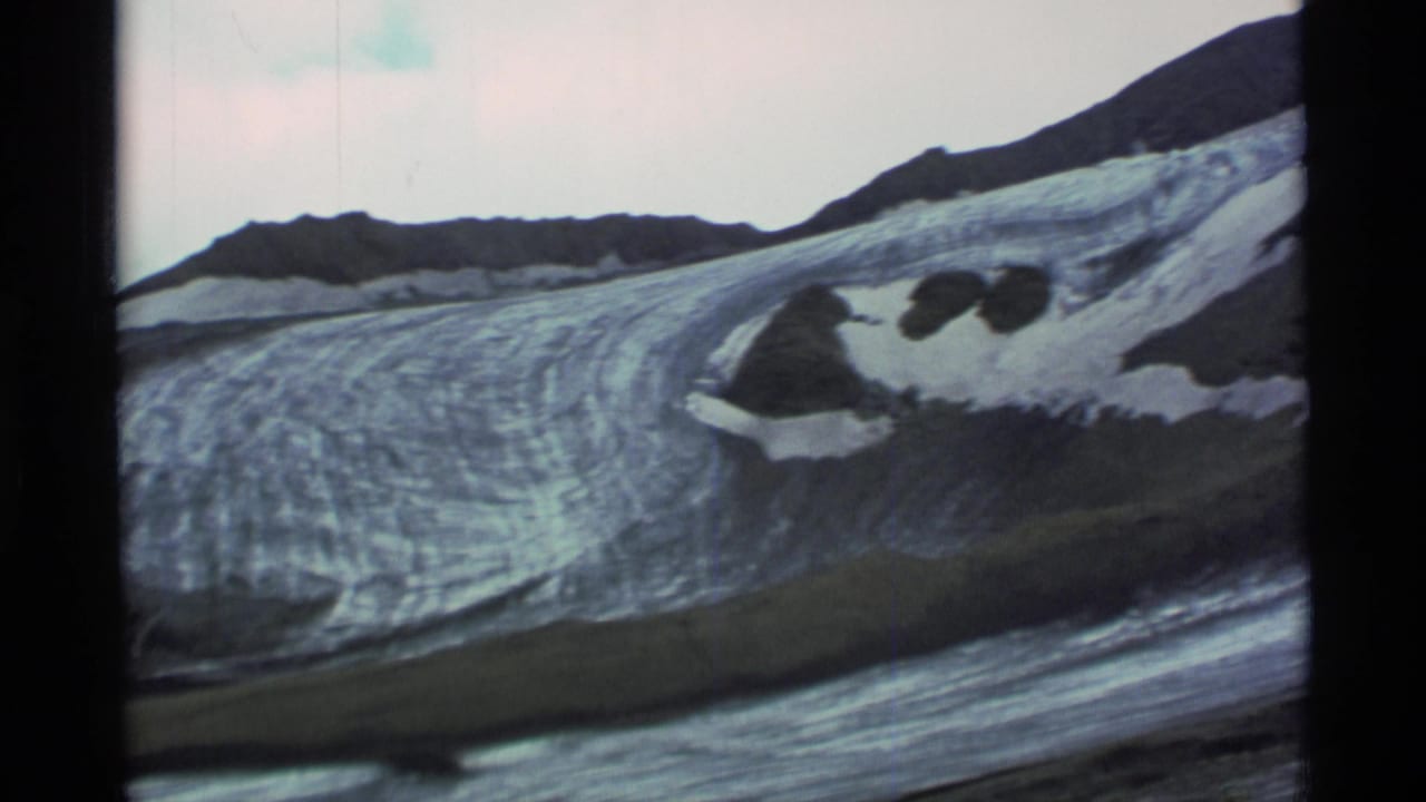 1977 Alaska home-movie frame of striated glacier ice running below dark mountains, with snow pockets and visible 8mm frame edges.