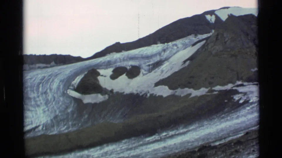 1977 Alaska 8mm frame showing glacier ice curving below a rugged mountain slope, with snow patches and dark rock visible.