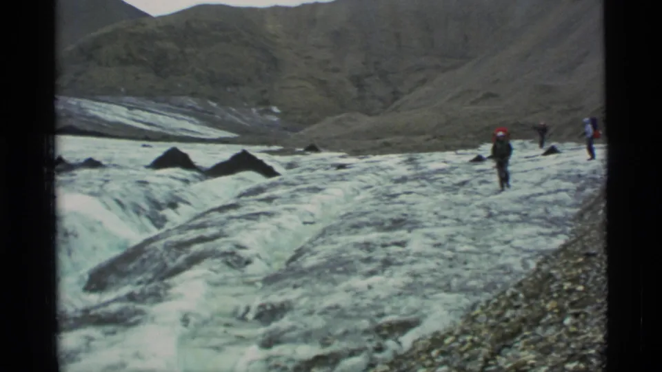 1977 Alaska home-movie frame showing distant hikers crossing glacier ice beside rocky terrain, with mountains rising in the background.