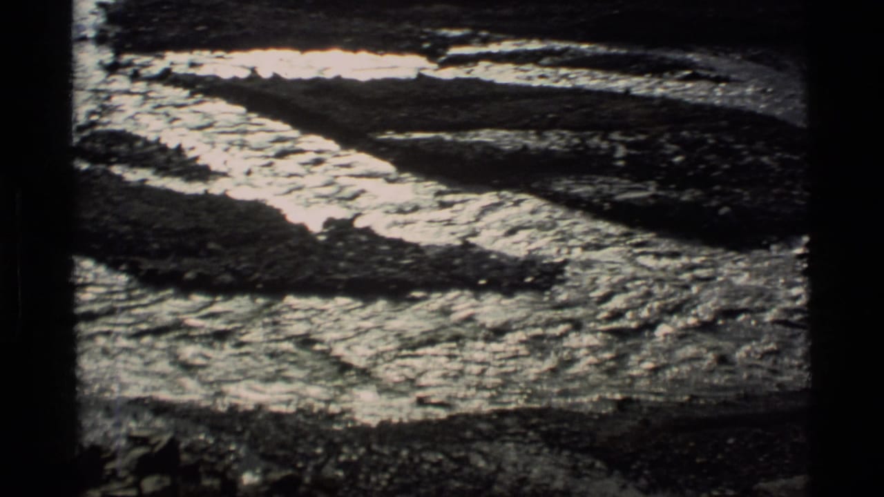 1977 Alaska 8mm frame showing reflective meltwater running through dark rocky ground with patches of snow or ice.