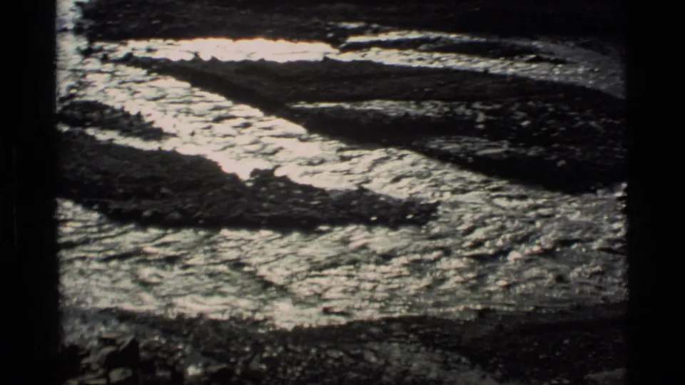 1977 Alaska 8mm frame showing reflective meltwater running through dark rocky ground with patches of snow or ice.