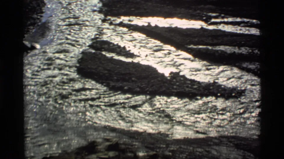 1977 Alaska home-movie frame with a narrow stream reflecting light as it passes between rocks and snow patches.