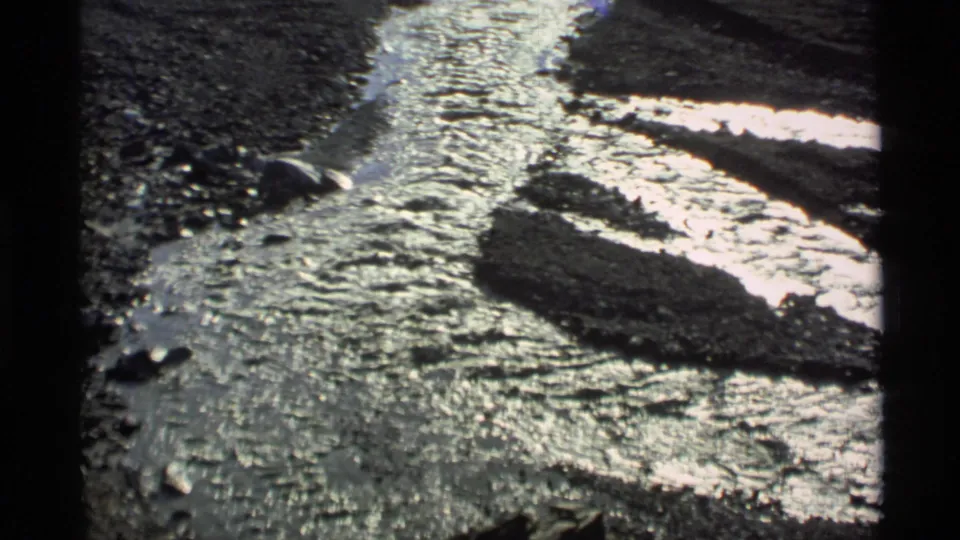 Vintage 8mm Alaska frame showing flowing meltwater, wet stones, and uneven snow or ice along a shallow channel.