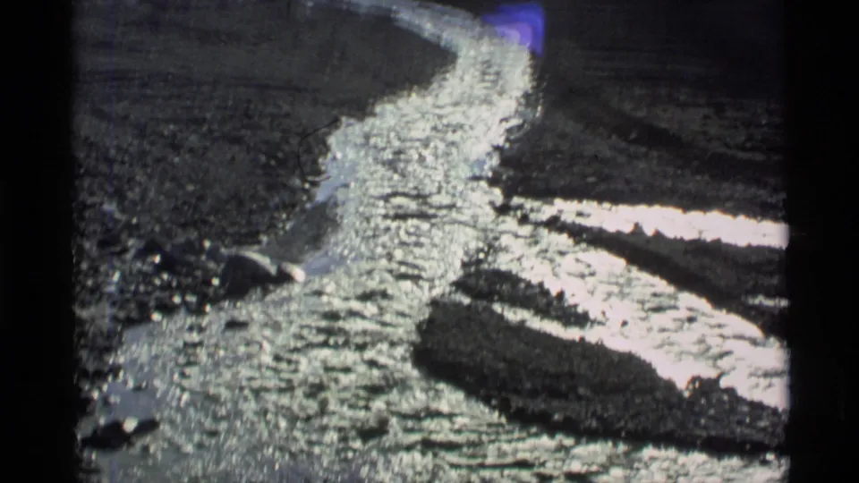 1977 Alaska 8mm frame showing a curving meltwater stream reflecting light beside dark rocks and remaining snow.