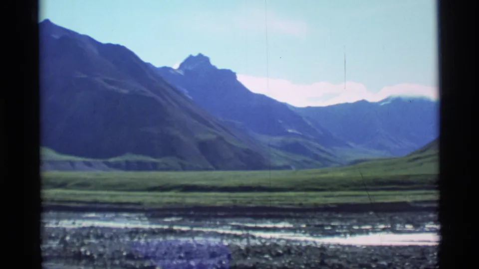 1977 Alaska 8mm frame showing a green open valley with blue mountain slopes and distant peaks below a bright sky.