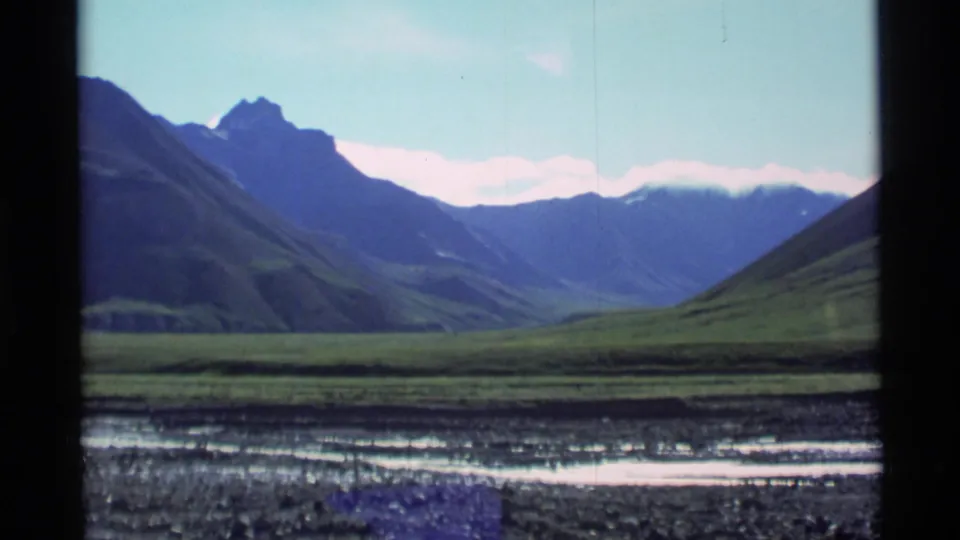 1977 Alaska 8mm frame showing a wide green valley framed by mountains, with distant peaks and bright sky beyond.