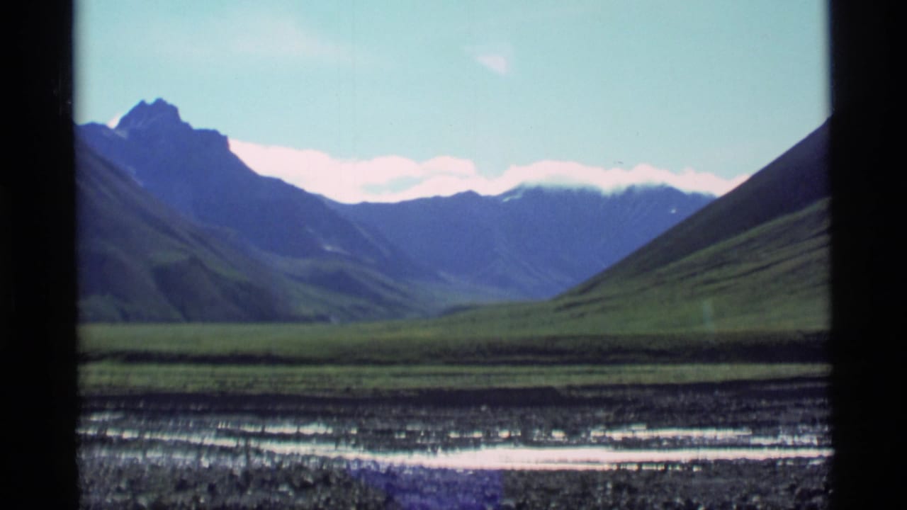 1977 Alaska 8mm frame showing a mountain valley through a window, with green hills, distant peaks, pale sky, and a rocky stream foreground.