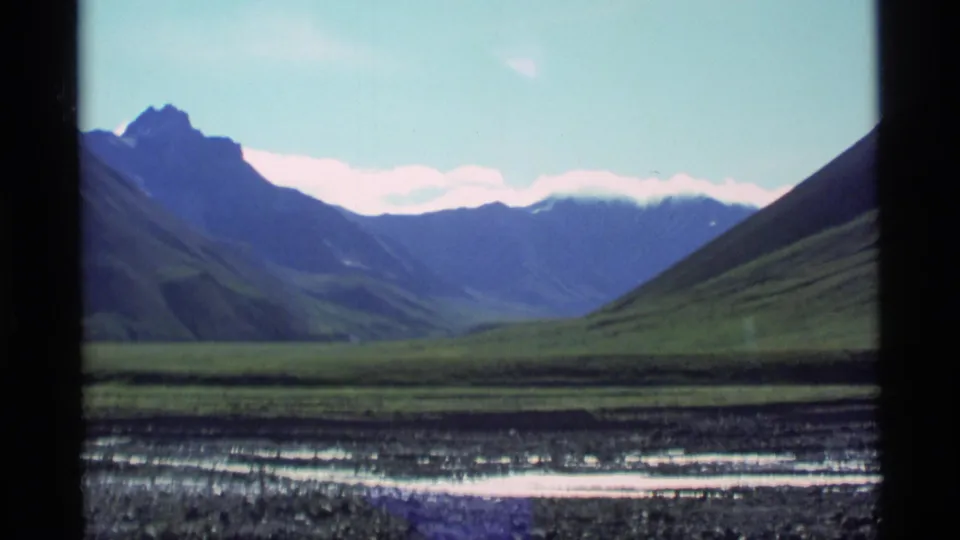 1977 Alaska 8mm frame showing a mountain valley through a window, with green hills, distant peaks, pale sky, and a rocky stream foreground.