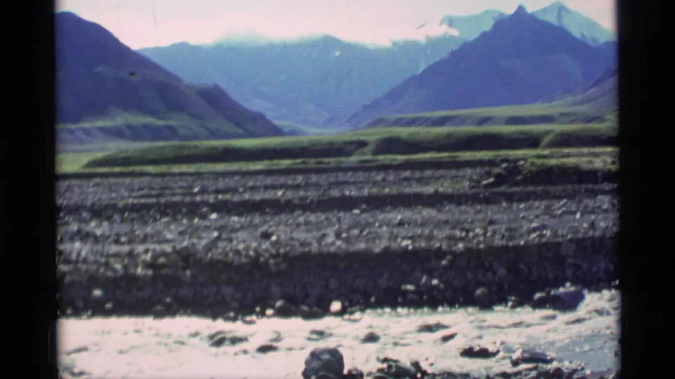 1977 Alaska 8mm frame of a broad valley beyond a rocky riverbed, with green terraces and blue mountains under a pale sky.