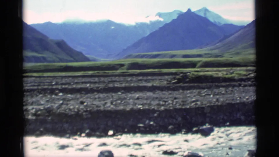 1977 Alaska 8mm frame with a moving stream and rocky bed in front of green valley landforms and distant mountain peaks.
