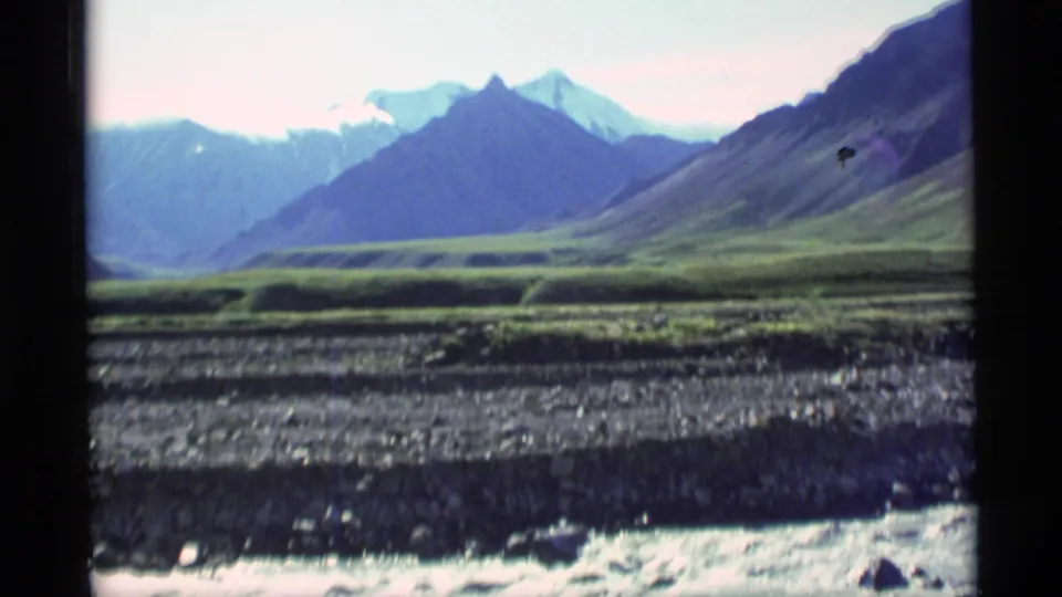1977 Alaska 8mm frame of steep mountain slopes, a green valley floor, rocky foreground, and pale sky seen through window edges.