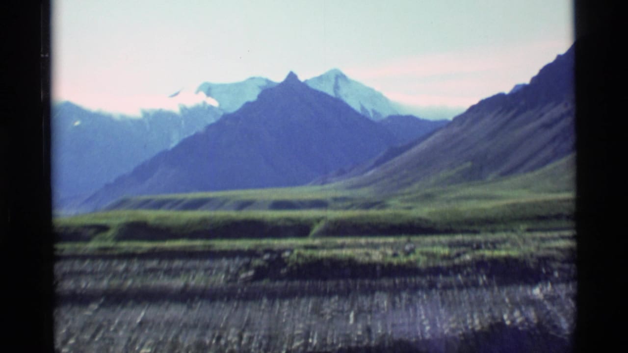 Distant snow-capped mountains above a green valley, seen within dark film-frame borders.