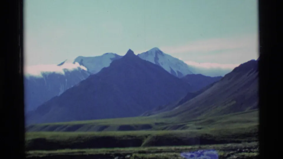 1977 Alaska 8mm frame of a mountain range beyond green open ground, with snow on distant peaks and a dark film-frame edge along the left side.