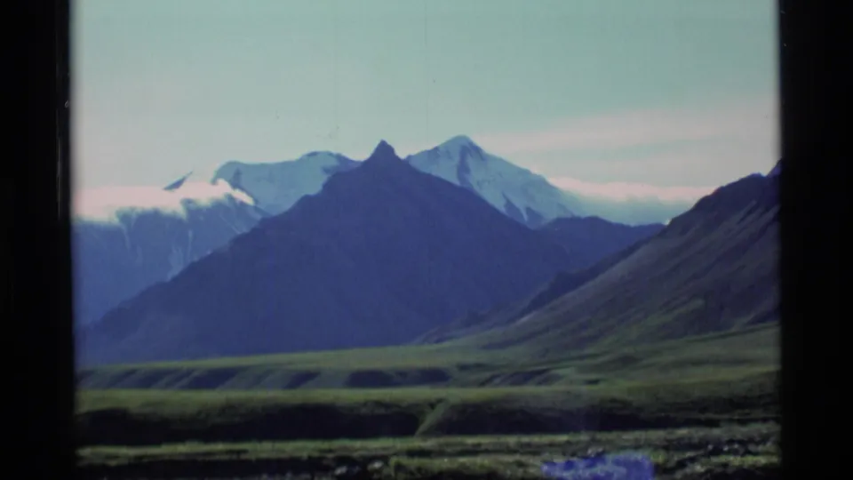 1977 Alaska 8mm frame showing a closer mountain view, with dark ridgelines in front of snow-capped peaks and green lower slopes.