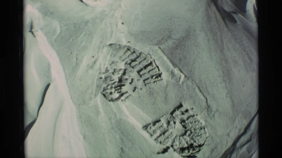 1977 Alaska 8mm frame showing boot prints and disturbed snow on a sloped snowy surface, adding a human trace without any visible person.