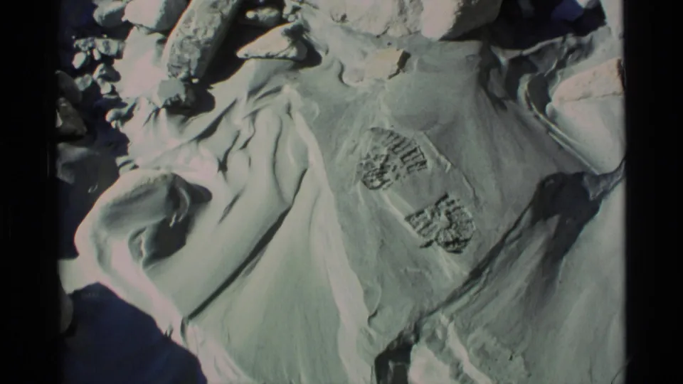 1977 Alaska home-movie frame showing snow, boot tread impressions, and surrounding rocky terrain.