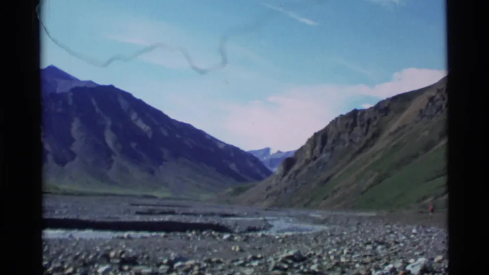 Wide 1977 Alaska 8mm landscape showing a rocky riverbed between steep green and dark mountain slopes.
