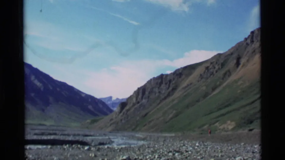 1977 Alaska 8mm frame of a remote mountain valley with a stony riverbed and blue sky.
