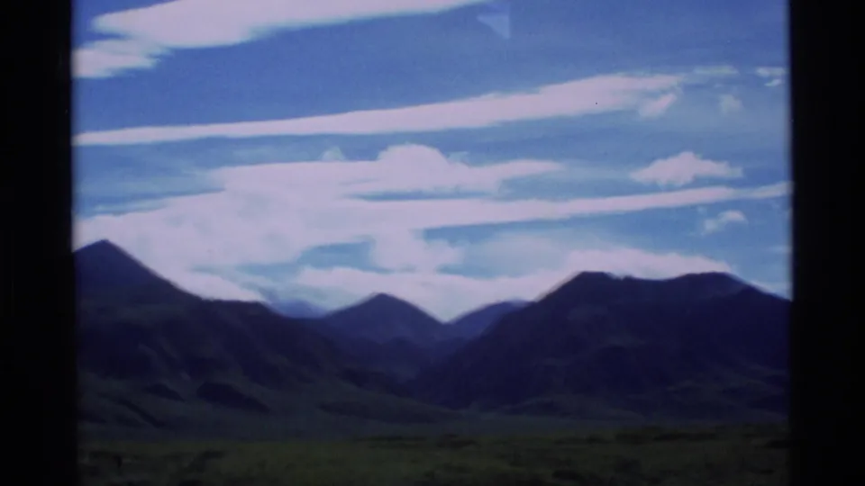 1977 Alaska 8mm frame showing dark mountain ridges beyond open grassland beneath a blue sky with layered white clouds.