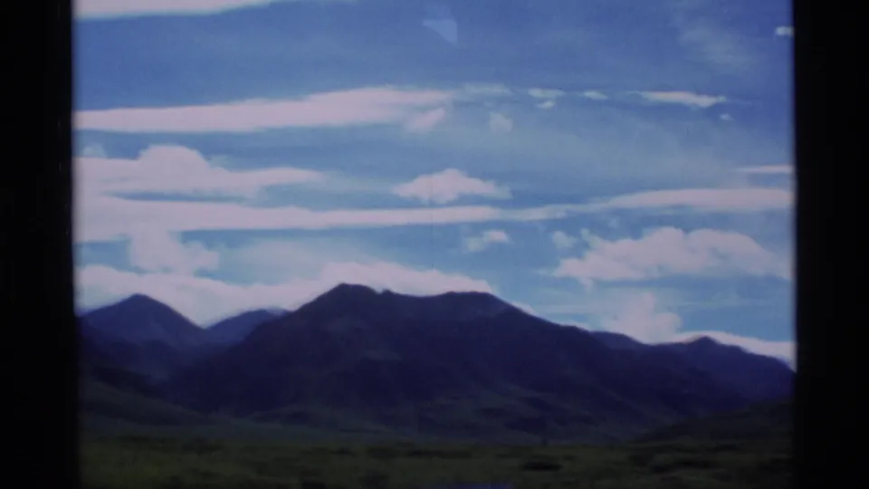 1977 Alaska home-movie view of a broad mountain skyline, green foreground, and bright cloud bands across a blue sky.