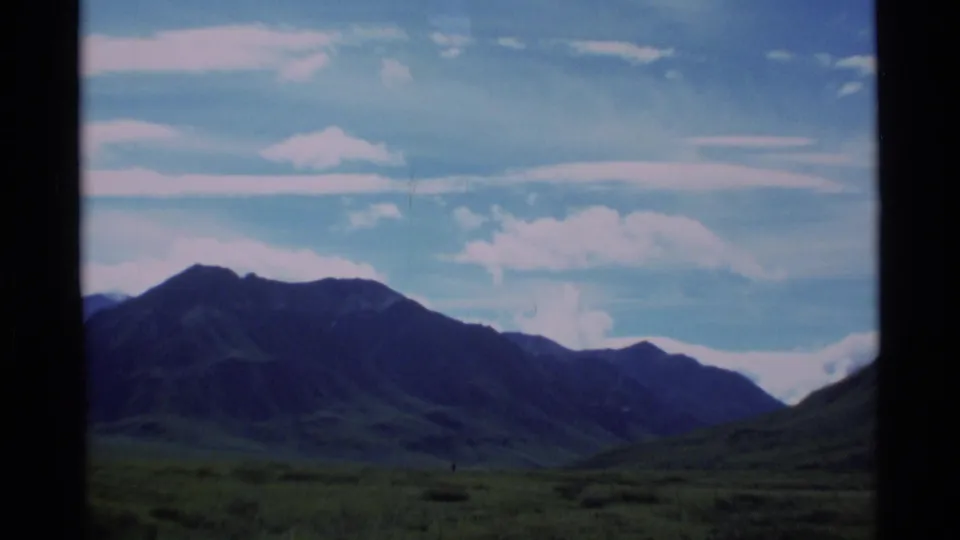 Soft 8mm Alaska landscape frame with a dark mountain slope rising over low grassland and pale clouds in a blue sky.