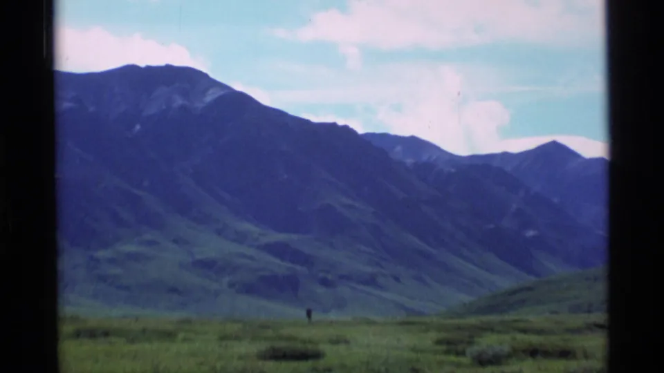 1977 Alaska 8mm frame showing a large mountain slope, grassy plain, bright clouds, and a tiny distant person for scale.