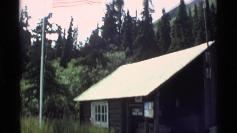 1977 Alaska home-movie frame showing a cabin roof and flagpole in front of tall evergreens and mountain-side vegetation.