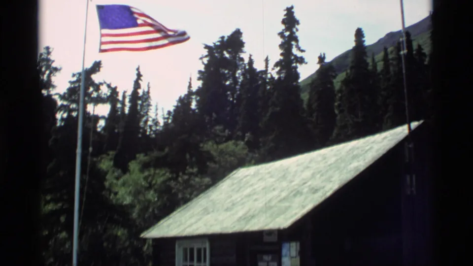 1977 Alaska 8mm frame with an American flag on a pole above a rustic cabin roof, backed by tall conifers and distant hillside.