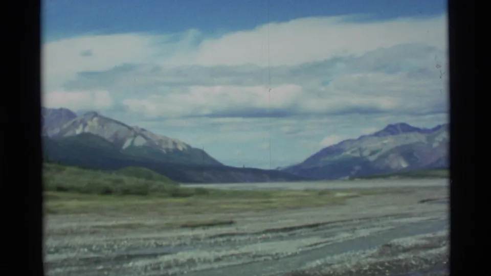 Wide 1977 Alaska landscape frame showing a flat rocky riverbed or plain, distant mountains, and layered clouds in a pale blue sky.