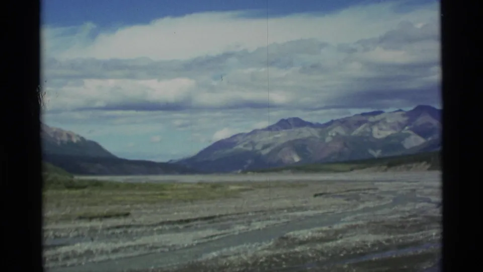 1977 Alaska home-movie frame with a broad valley, distant pass, and mountains beneath a bright clouded sky.