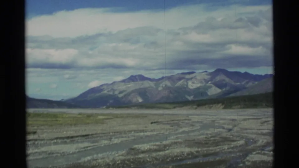 1977 Alaska 8mm frame showing gravel flats, low water channels, and a mountain range under heavy clouds.
