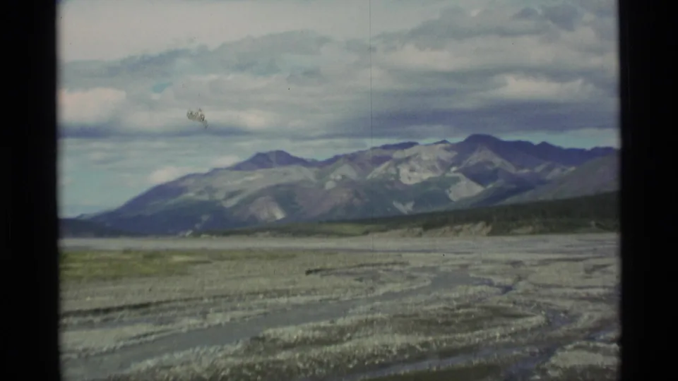 1977 Alaska home-movie frame with braided gravel terrain, distant mountains, and visible dark 8mm frame edges.