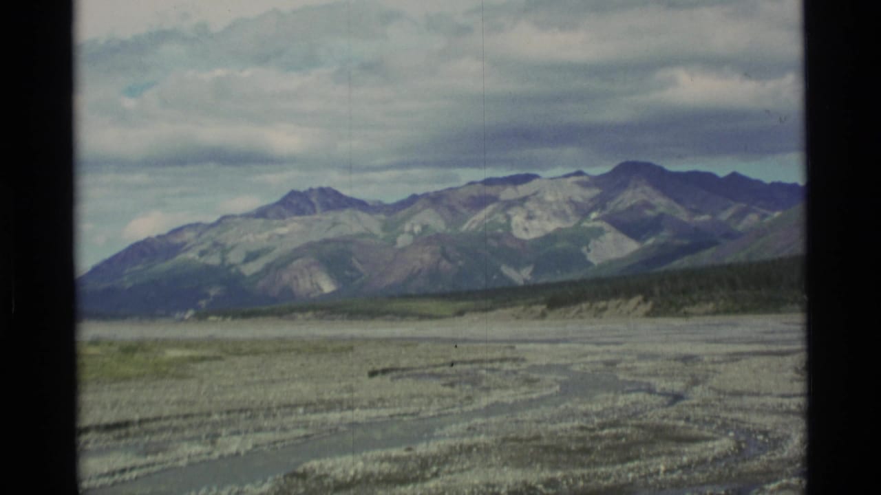 1977 Alaska 8mm frame showing distant rugged mountains, cloudy sky, and a broad pale riverbed or gravel wash.