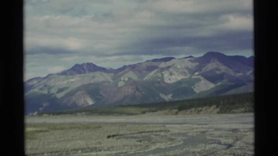 1977 Alaska landscape frame with mountain peaks, gray clouds, a broad gravel wash, and dark 8mm gate borders.