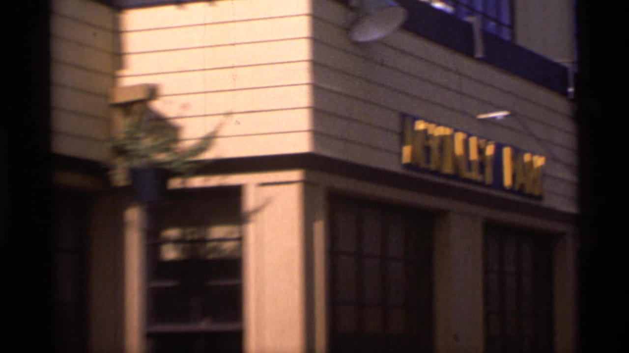 1977 Alaska home-movie frame showing a building corner with a yellow-lettered park-style sign and large windows.