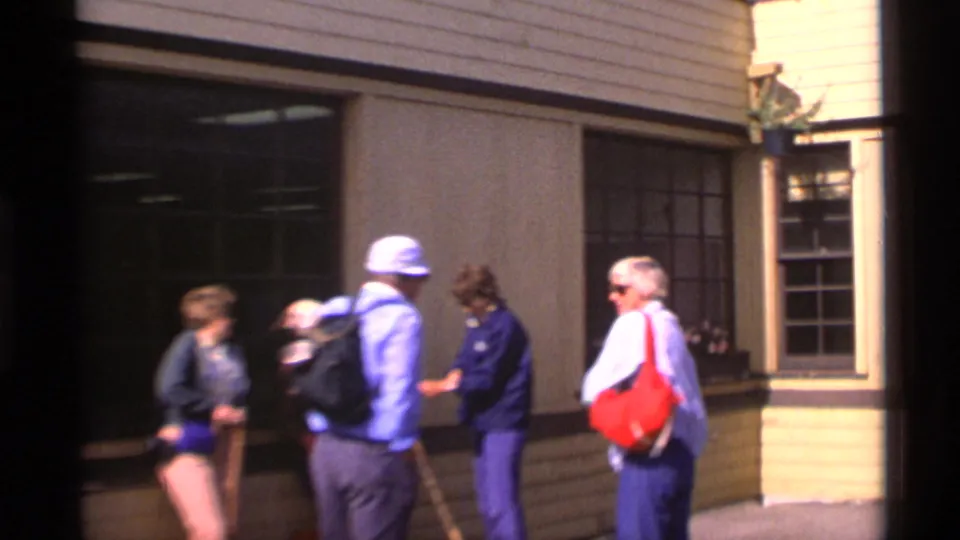 Four adult tourists in 1970s casual clothing gather outside an Alaska visitor building, with bags and walking sticks visible.