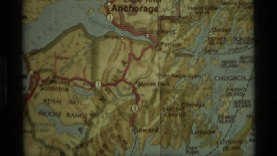 1977 home-movie frame of a folded Alaska map showing red-marked road routes around Anchorage, Seward, and Kenai Peninsula.