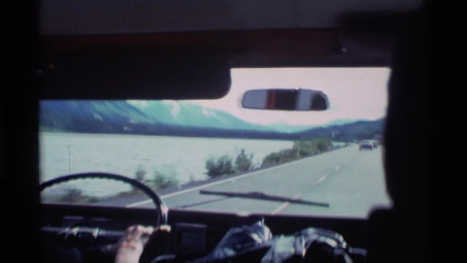 1977 Alaska road-trip POV from inside a vehicle, looking past the windshield toward a lakeside or coastal road and snowy mountains.