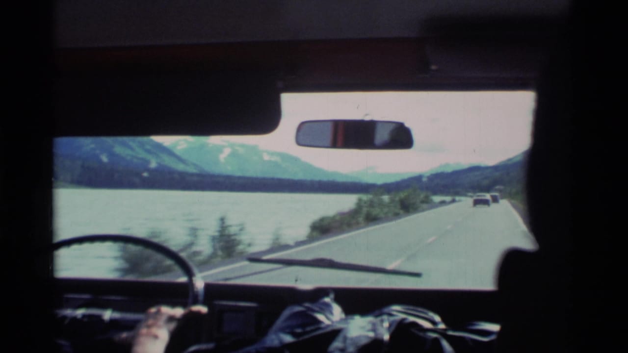 View through a car windshield of a coastal road beside water and mountains.