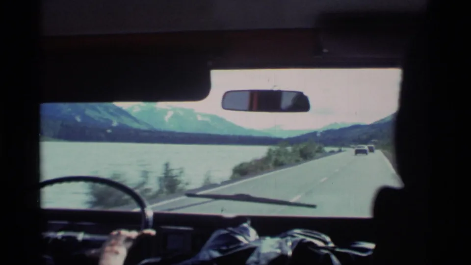 1977 Alaska 8mm frame from inside a car, showing a driver’s hand, windshield, shoreline road, water, and distant mountains.