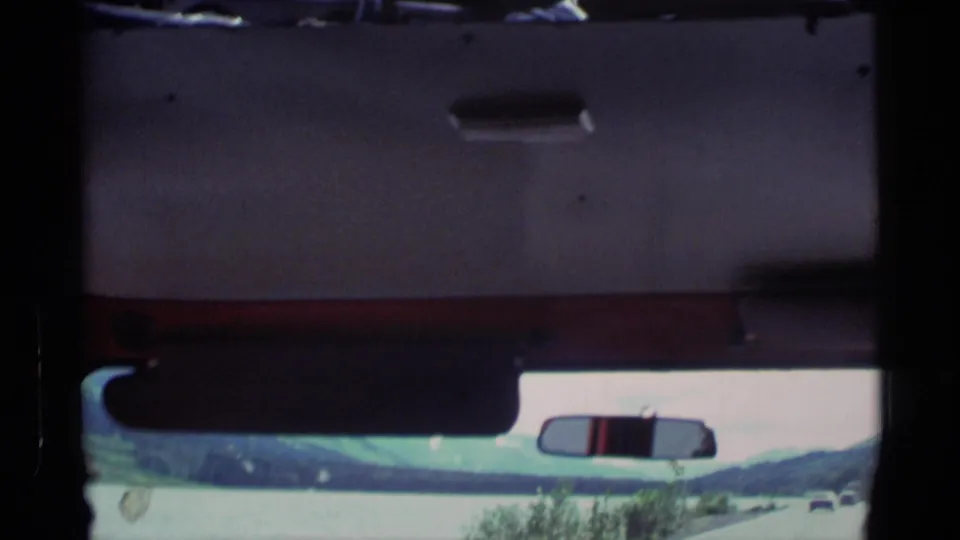 1977 Alaska road-trip frame with the windshield and rearview mirror framing a coastal road beside water and mountain slopes.