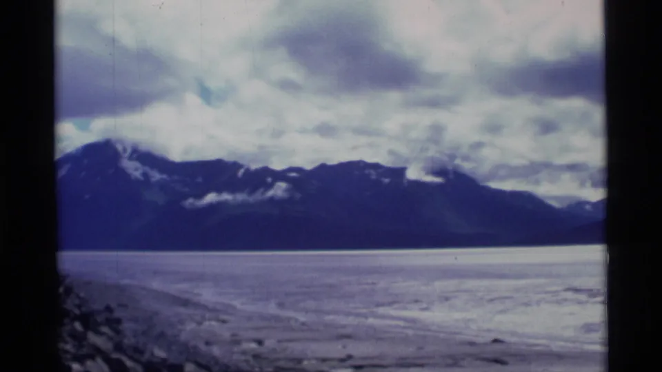 1977 Alaska 8mm landscape frame showing broad water, a low shoreline, dark mountain range, and heavy clouds.