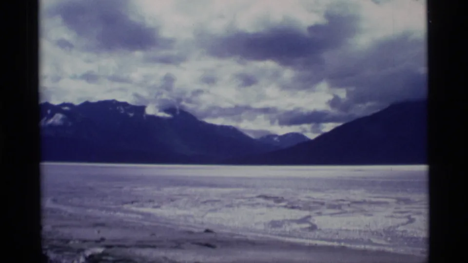 1977 Alaska 8mm frame showing a broad pale flatland or icy surface below dark mountain ridges and clouded sky.