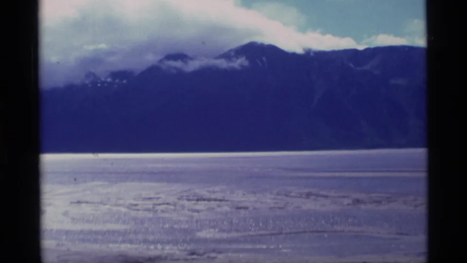 1977 Alaska home-movie frame with a dark mountain wall, bright flat foreground, and low clouds moving across the peaks.